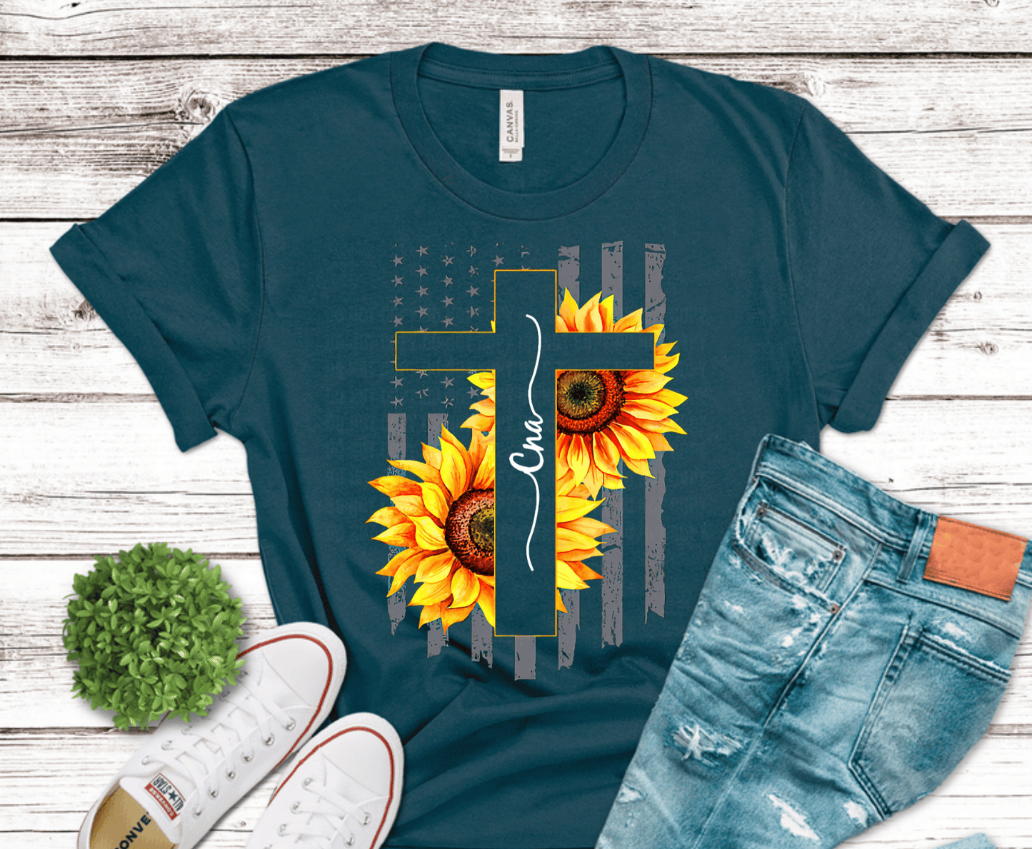 Teal t-shirt with sunflower and cross design, paired with jeans and sneakers on a wooden surface.