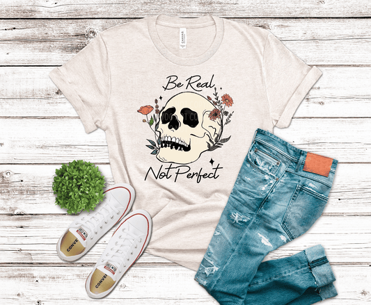 Floral skull graphic with be real not perfect text surrounded by flowers and artistic elements