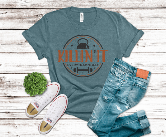 Teal t-shirt with 'KILLIN' IT EVERY DAMN DAY' graphic, jeans, and sneakers on a wooden surface.