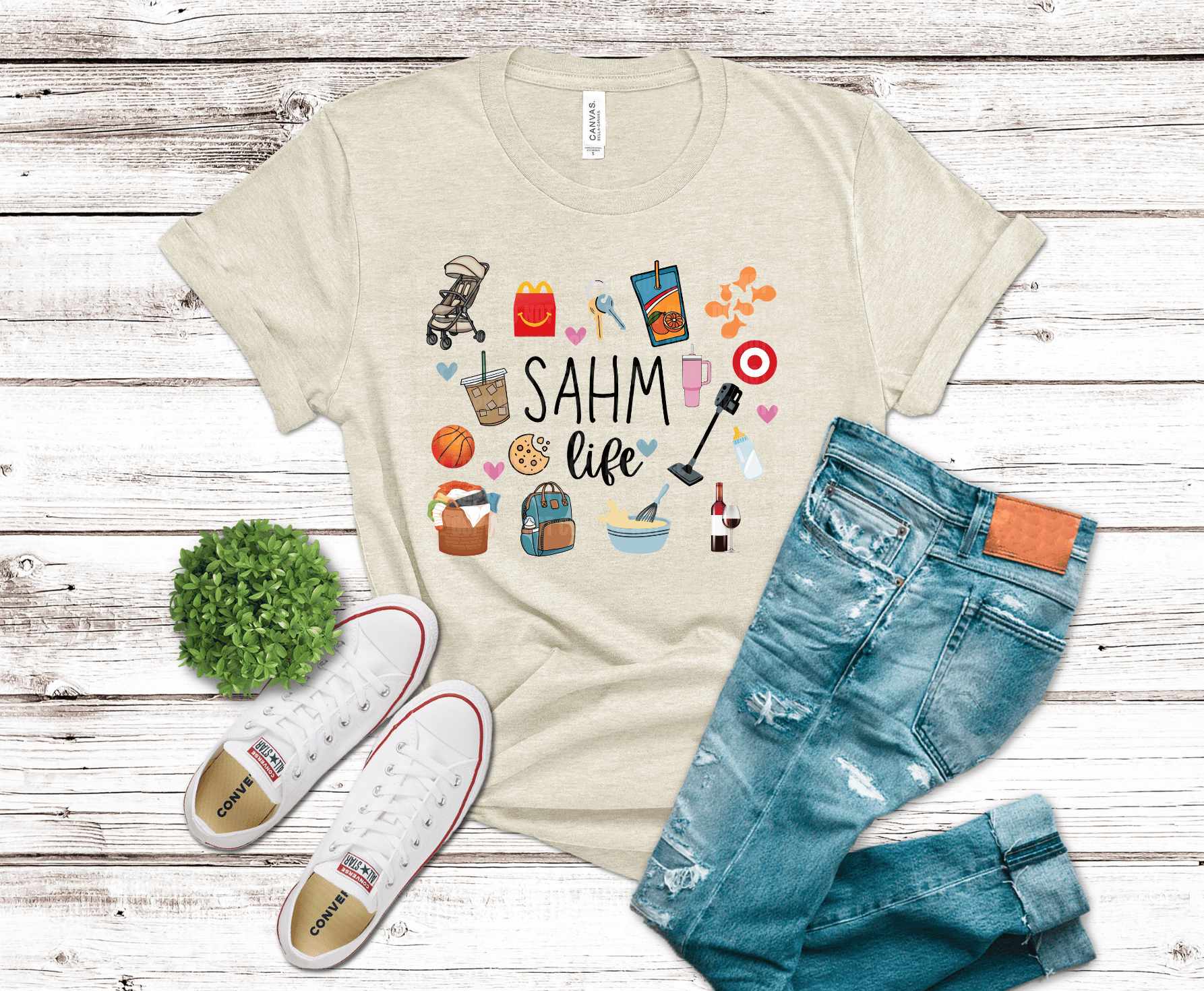 Beige t-shirt with 'SAHM life' graphic, blue jeans, and white sneakers on a wooden surface