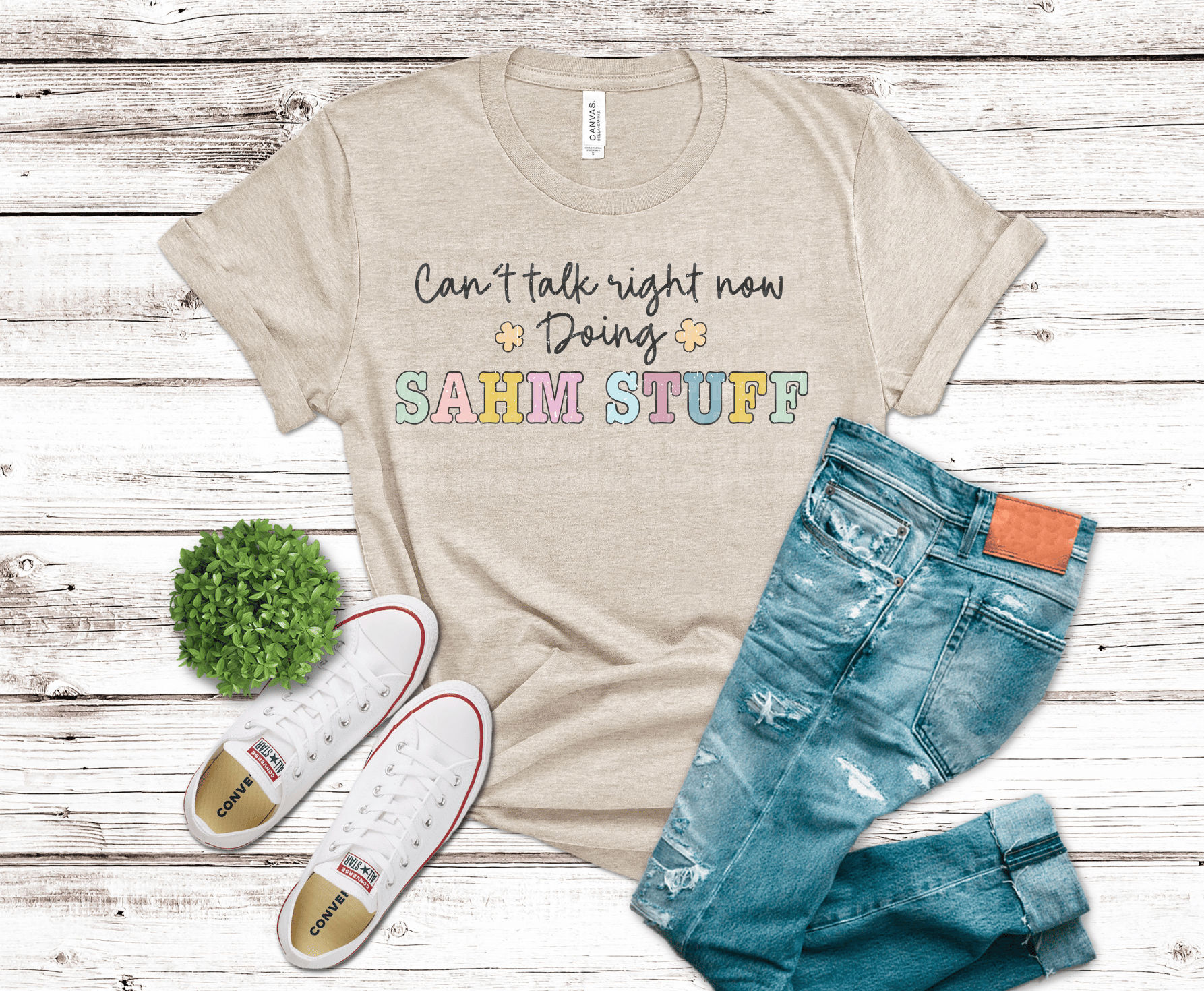 Beige t-shirt with colorful text, blue jeans, and white sneakers on a wooden surface