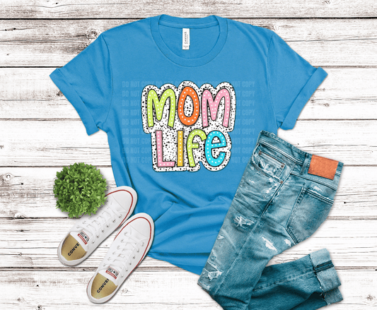Blue t-shirt with 'Mom Life' graphic, jeans, and sneakers on a wooden surface